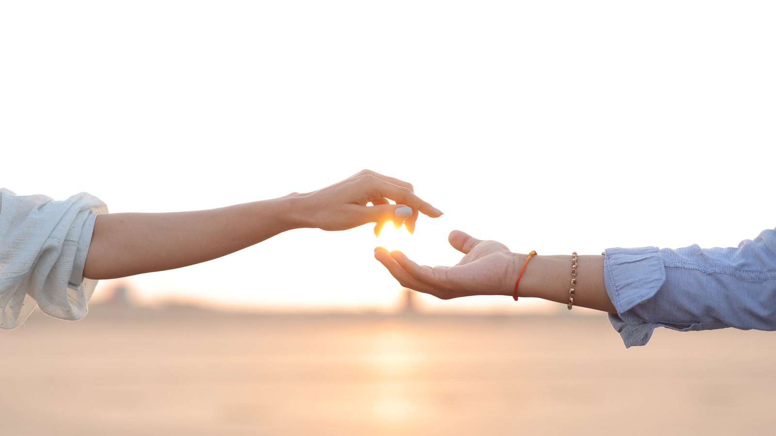 Two arms reaching toward each other against a bright sunset, fingertips almost touching, with the light glowing in the gap between them.